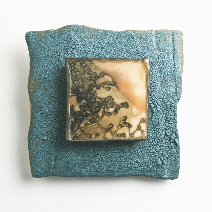 Small, blue ceramic tile with an eco print  of  botanical seed pods encased in wax and mounted in the center. Artwork by Sarah Treanor available at larkandkey.com.