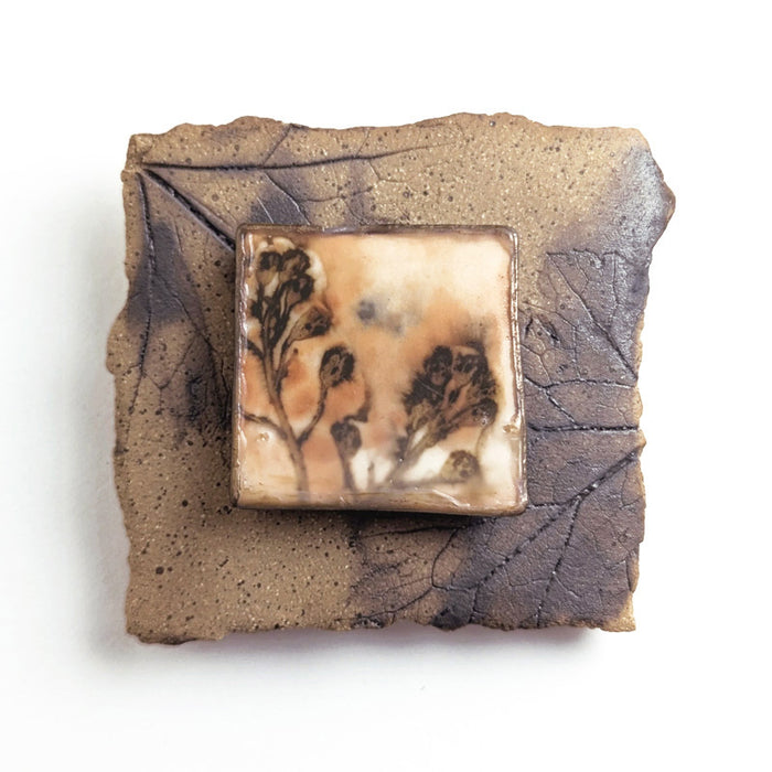 Small ceramic tile with textured blue glaze, with a botanical print encased in wax set in the center. Artwork by Sarah Treanor. 