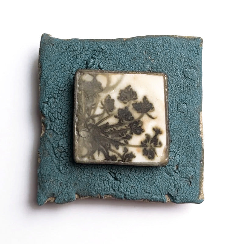 Small ceramic tile with textured blue glaze, with a botanical print encased in wax set in the center. Artwork by Sarah Treanor. 