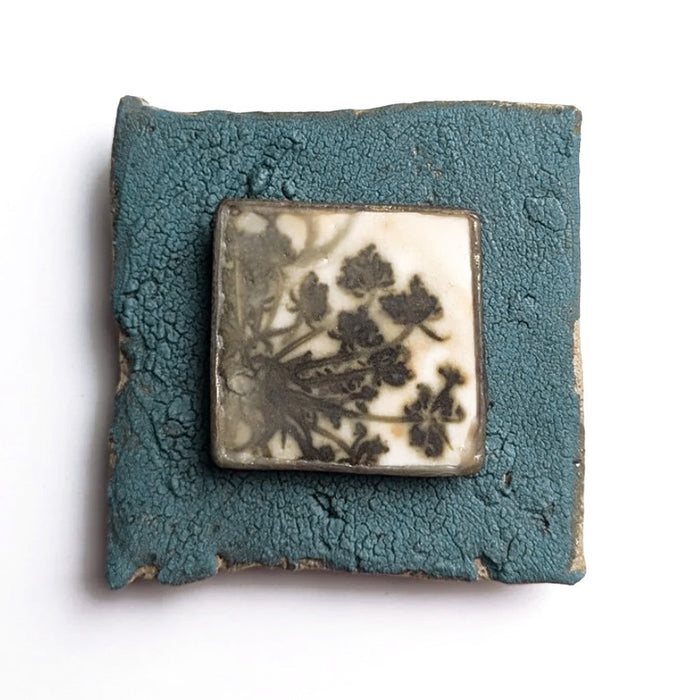 Small ceramic tile with textured blue glaze, with a botanical print encased in wax set in the center. Artwork by Sarah Treanor. 