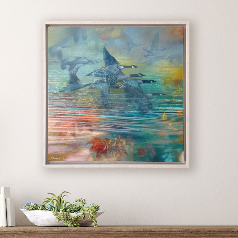 Oil painting with geese flying over reflective water in soft pastel tones of blue, teal, green, yellow, and orange. The artwork, by Sharon Cosgrove, is set in a hardwood maple frame and displayed on a white wall over a table. 