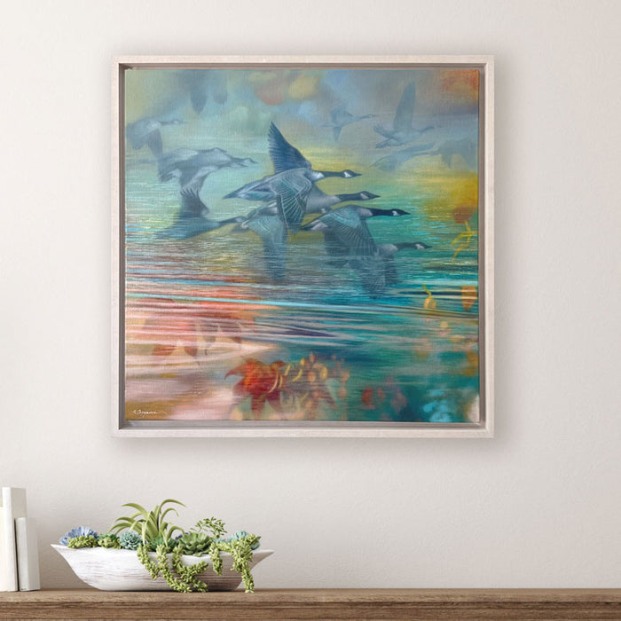 Oil painting with geese flying over reflective water in soft pastel tones of blue, teal, green, yellow, and orange. The artwork, by Sharon Cosgrove, is set in a hardwood maple frame and displayed on a white wall over a table. 