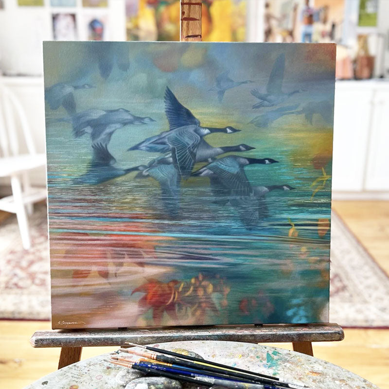 Oil painting with geese flying over reflective water in soft pastel tones of blue, teal, green, yellow, and orange. The artwork, by Sharon Cosgrove, is shown in the artist's studio. 