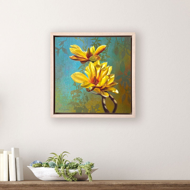 Oil painting featuring two bright yellow magnolia flowers  against a textured blue and green botanical background. The artwork, by Sharon Cosgrove, is set in a hardwood maple frame and displayed over a table.