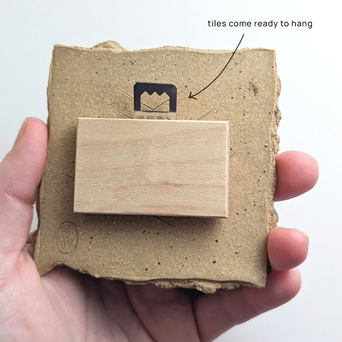 Hand holding small ceramic tile, showing hanger on back. 
